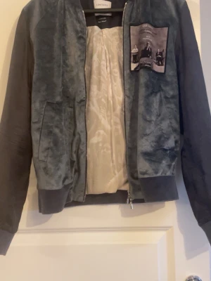 Mörkgrå bomberjacka från Limitato - Snygg bomberjacka från Limitato med sammetsliknande material på framsidan och släta ärmar. Jackan har en unik patch med foto på bröstet och ljus innerfoder med mönster. Dragkedja framtill och ribbade muddar vid ärmslut och midja. Liten slitning på armen som knappt syns. Passar XS också