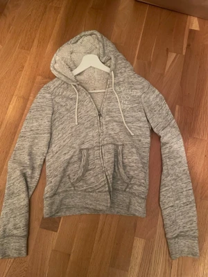 Grå zip hoodie från Hollister  - Mysig gråmelerad hoodie från Hollister med dragkedja, broderad logga och text på bröstet. Utrustad med huva och vita snören samt två fickor framtill. Insidan är fodrad med mjukt teddyfoder för extra komfort. Perfekt för chill dagar. Skriv för fler bilder eller frågor! Pris kan vara diskuterbart☺️