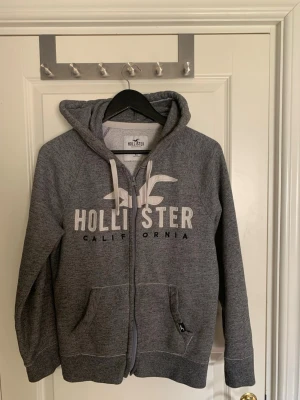Hollister zip  - Snygg grå hoodie från Hollister med dragkedja,passar dig som har S vanligt vis eller liten M.  stor logga och texten 'California' på bröstet. Mjuk bomullsblandning, huva med vita snören och två fickor framtill. Perfekt för chill dagar eller till skolan.