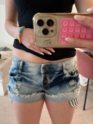 Blå slitna jeansshorts med ränder - Säljer ett par blå denimshorts med slitna detaljer och uppvikta ben. Shortsen har knappar framtill och coola vit- och grårandiga detaljer på insidan av benen och fickorna. Perfekta för sommaren och ger en avslappnad streetstyle-vibe. Köpta på Vintage&Friends men märket är rainbow