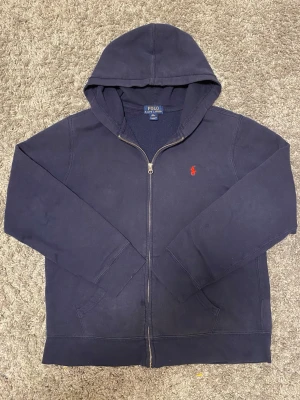 Ralph Lauren zip  - Mörkblå hoodie med dragkedja från Polo Ralph Lauren. Klassisk design med huva och fickor framtill, samt det ikoniska röda Polo-logot broderat på bröstet. Materialet är mjukt och bekvämt, perfekt för en avslappnad look.