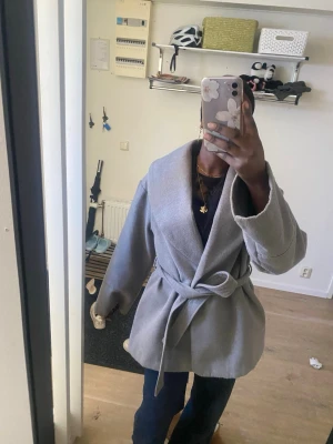 Grå omlottkappa med knytskärp - Snygg grå kappa i omlottmodell med brett knytskärp i midjan. Kappan har bred krage och raka ärmar, vilket ger en avslappnad men stilren look. Perfekt att slänga över en hoodie eller stickad tröja. Materialet ser ut att vara mjukt och lite tjockare tyg.