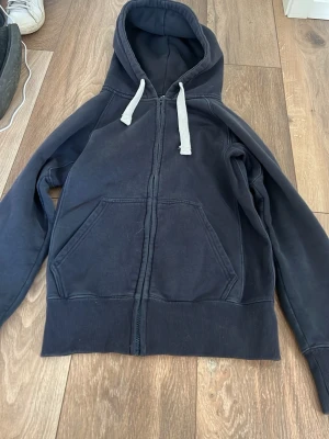 Svart zip-hoodie med vita snören XS - En svart hoodie med dragkedja från CR, storlek XS. Den har vita snören i huvan och två stora fickor framtill. Mjuk insida och ribbade muddar vid ärmslut och nederkant. Perfekt för en chill och avslappnad stil.