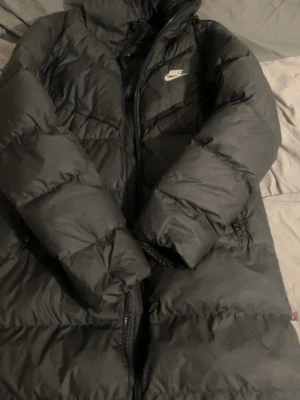 Svart lång dunjacka från Nike - Säljer en svart lång dunjacka från Nike med klassisk logga på bröstet. Jackan har dragkedja framtill, hög krage och långa ärmar. Perfekt för kalla dagar med sin tjocka vaddering och stilrena look.