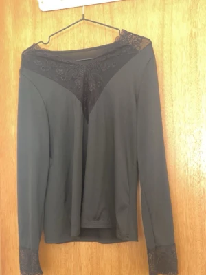 Svart långärmad topp med spets - Svart långärmad topp med bred spetsdetalj över axlar och halsringning samt spets vid ärmslut. Toppens material är mjukt och stretchigt, vilket ger en bekväm passform. Perfekt för dig som gillar en stilren look med lite extra detaljer.