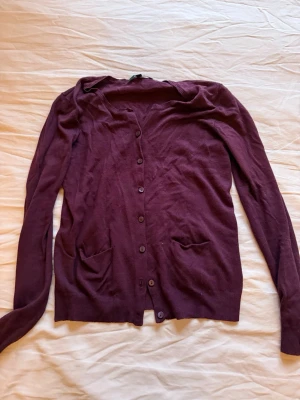 Vinröd kofta med knappar - Vinröd kofta med långa ärmar och knäppning framtill. Två små fickor framtill och ribbade muddar vid ärmslut och nederkant. Perfekt att slänga över en t-shirt eller linne för en chill look.
