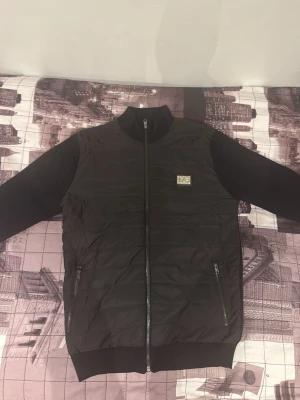 Svart bomberjacka med dragkedja - Snygg svart bomberjacka med hel dragkedja framtill och ribbade muddar vid ärmar och nederkant. Jackan har två fickor med dragkedja och en liten patch på bröstet. Perfekt för en stilren och avslappnad look.