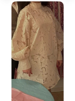 Ljus kort klänning med volangärm - Säljer en ljus kort klänning med hög hals och voluminösa långärmar. Klänningen har ett genombrutet mönster och är tillverkad i ett tunt, strukturerat tyg som ger en elegant look. Perfekt för dig som gillar unika detaljer och en loose passform.