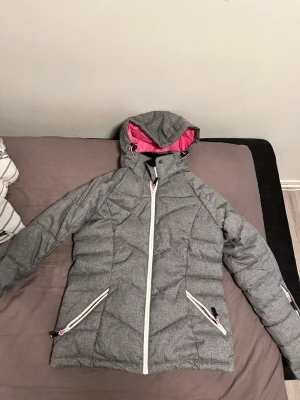 Grå vindjacka från Tuxer XS - Snygg grå vindjacka från Tuxer med vit dragkedja och detaljer. Jackan har huva med rosa foder, två snedställda fickor med dragkedja och sportig design. Perfekt för blåsiga dagar och outdoor-aktiviteter.