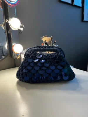 Mörkblå kuvertväska med paljetter - Engenvirkad mörkblå kuvertväska med stora glansiga paljetter. Väskan har ett inbyggt handtag upptill och en mjuk, lite puffig form. Perfekt för dig som vill sticka ut med accessoarerna och gillar en unik look.