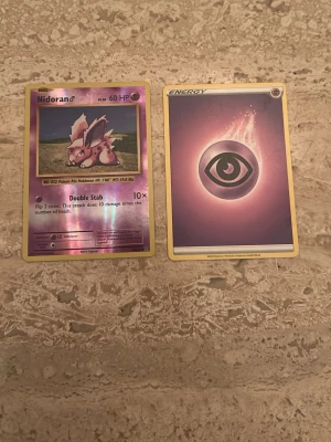 Pokémonkort Nidoran och Psychic Energy - Två Pokémonkort: ett holografiskt Nidoran-kort med lila och rosa färger samt ett Psychic Energy-kort med svart symbol på lila bakgrund. Båda korten har klassisk Pokémon-design och är perfekta för samlare eller spelare.