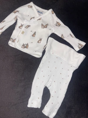 Vitt babyset från Rikiki med kaniner - Supersött tvådelat babyset från Rikiki i storlek 50. Setet består av en vit omlottbody med långa ärmar och gulligt kaninmönster samt matchande byxor med små prickar. Tillverkat i mjukt ribbat tyg som är skönt för de allra minsta.