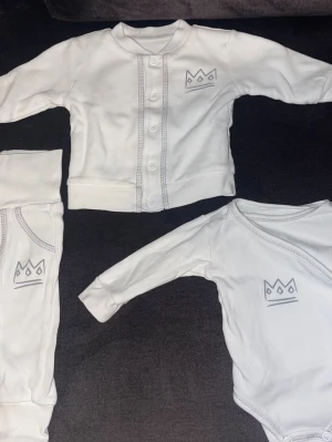 Vitt babyset med kronmotiv - Sött vitt set för baby med långärmad body, byxa och kofta. Alla delar har ett stilrent kronmotiv tryckt på bröstet eller benet. Setet är tillverkat i mjukt och bekvämt tyg, perfekt för små barn. Koftan har knäppning framtill och plaggen har diskreta sömmar.