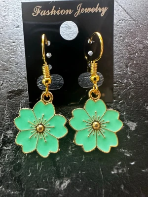Gröna blomörhängen med gulddetaljer - Snygga örhängen i form av gröna blommor med guldfärgade kanter och detaljer. Hängande modell med krok, perfekta för att ge din look en färgklick. Materialet är metall och emalj.