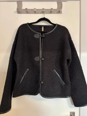 Svart cardiganjacka från Soyaconcept - Stilren svart cardiganjacka från Soyaconcept med snygga detaljer i fuskskinn längs kanterna och knäppning framtill. Jackan har två fickor med fuskskinnskanter och är tillverkad i ett mjukt, strukturerat material. Perfekt för lager-på-lager-stil eller en tunn jacka för höst/vår. 