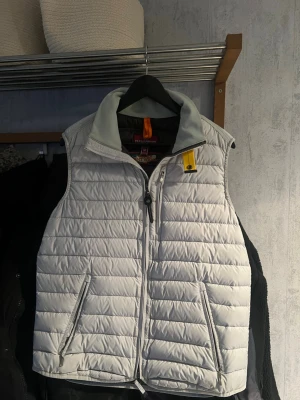 Ljusgrå dunväst från Parajumpers - Snygg ljusgrå dunväst från Parajumpers med ståkrage och dragkedja framtill. Västen har två dragkedjeförsedda fickor och en gul detalj vid halsen. Tillverkad i superlätt material som håller dig varm utan att kännas tung.