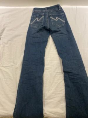 Blå bootcut jeans från Nelly - Snygga blå bootcut jeans från Nelly med dubbla knappar i midjan och dragkedja. Jeansen har klassiska bakfickor med dekorativa sömmar och är tillverkade i ett mjukt denimtyg. Perfekta för en trendig och avslappnad look.