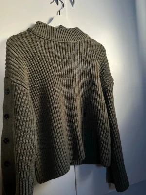 Olivgrön stickad tröja med knappar - Snygg olivgrön stickad tröja med grov ribbstickning och svarta knappar längs ärmarna. Tröjan har en hög krage och avslappnad passform, perfekt för lager-på-lager. Materialet känns mjukt och värmande.