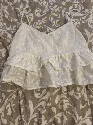 Vit broderad linnetopp med volang - Superfin vit linnetopp med tunna axelband och broderade blommor. Toppen har volangdetalj och är croppad, perfekt för varma dagar. Söt och somrig stil som passar till jeans eller kjol. Storlek S. 