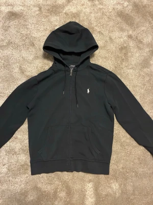 Svart zip hoodie från Ralph Lauren - Svart hoodie med dragkedja från Polo Ralph Lauren. Klassisk design med huva och dragsko, två fickor framtill och den ikoniska vita logotypen broderad på bröstet. Perfekt för en avslappnad och stilren look.