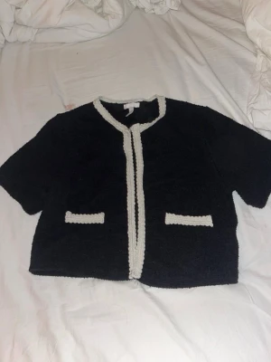 Svart kofta med vita detaljer - Kort svart kofta med raka, halvlånga ärmar och dekorativa vita kanter längs öppningen och fickorna. Koftan har två fejkfickor framtill och är gjord i ett mjukt, stickat material. Perfekt för en chic och enkel look. Vet inte helt för hur mycket men säljer för bara 150 kr!!!! Köp köp köp!