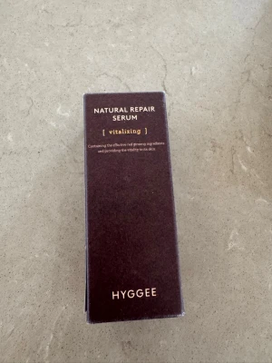 Hyggee Natural Repair Serum - Vitaliserande serum från Hyggee med röd ginseng, perfekt för att ge huden extra lyster och näring. Kommer i en mörklila rektangulär förpackning med guldfärgad text. Innehåller 30 ml och passar för dig som vill boosta din hudvårdsrutin.