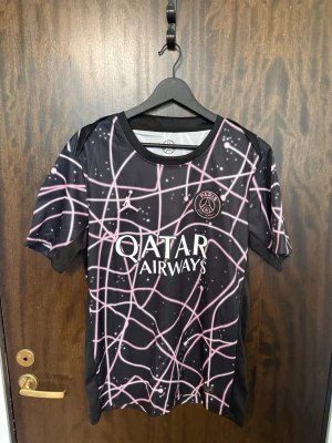 PSG x Jordan svart matchtröja - Svart PSG matchtröja i samarbete med Jordan, med rosa och vita grafiska linjer över hela tröjan. Tryckt Paris Saint-Germain-logga på bröstet, Jordan-logga och Qatar Airways som sponsor. Tillverkad i lätt och ventilerande Dri-FIT-material.