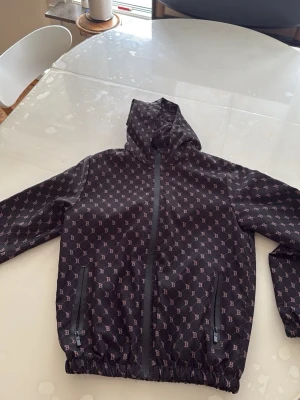 Svart vindjacka med mönster från burberry - Snygg svart vindjacka från Burberry med heltryckt mönster av små B-loggor i ljusrosa. Jackan har huva, dragkedja framtill och två fickor med dragkedja. Resår i midja och ärmslut ger en schysst passform. Perfekt för dig som gillar streetstyle och designerplagg.