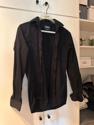 Svart skjorta från Dressmann - Klassisk svart skjorta från Dressmann med lång ärm och knappar framtill. Skjortan har en stilren design med normal passform och är tillverkad i bomull, perfekt för en clean och enkel look.