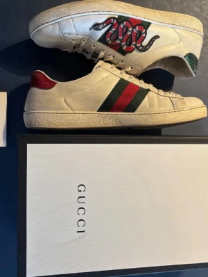 Gucci Ace  - Gucci Ace sneakers i vitt läder med klassiska gröna och röda sidoremsor samt broderad orm i rött och svart på sidan. Baksidan har glitterdetaljer i rött och grönt. Snygg och ikonisk modell som sticker ut.