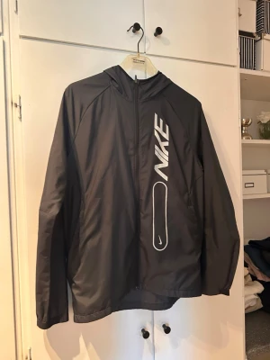 Svart vindjacka från Nike med huva - Snygg svart vindjacka från Nike med dragkedja och huva. Jackan har en stor vit Nike-logga på framsidan och reflekterande detalj bak. Lätt material som passar perfekt för blåsiga dagar. Sportig och stilren design.