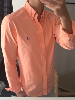 Persikofärgad skjorta från Ralph Lauren - Persikofärgad skjorta från Ralph Lauren med klassisk button-down krage och blå broderad logga på bröstet. Tillverkad i mjuk bomull och har långa ärmar samt knappar framtill. Perfekt för en clean och stilren look. Den är ganska urtvättad, därav priset. Annars bra skick 