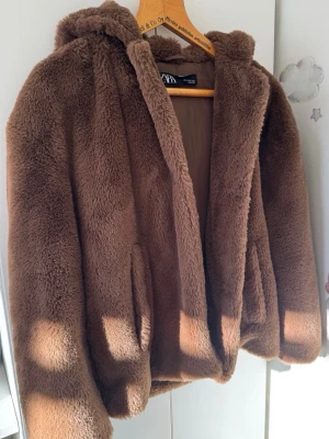 Brun teddyjacka med huva från ZARA - Supermjuk och fluffig brun teddyjacka från ZARA med huva och två fickor framtill. Jackan har en avslappnad passform och är fodrad på insidan för extra komfort. Perfekt för kyliga dagar när du vill vara både varm och snygg.