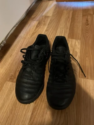 Svarta Nike fotbollsskor - Säljer ett par helsvarta fotbollsskor från Nike med klassisk låg profil och snörning. Skorna har en ovandel i syntetmaterial och gummisula med små dobbar, perfekt för spel på konstgräs eller grus. Snygg och stilren design med diskret Nike-logga på sidan.