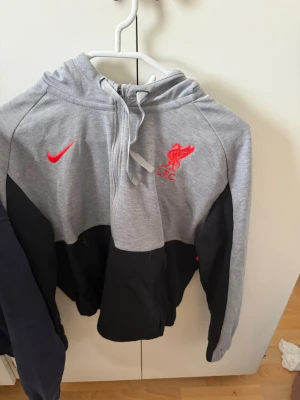 Grå och svart LFC Nike hoodie - Snygg hoodie från Nike i grått och svart med röd LFC-logga och Nike swoosh på bröstet. Tröjan har huva med dragsko och en röd detalj på ryggen. Perfekt för dig som gillar fotboll och vill ha en sportig stil.