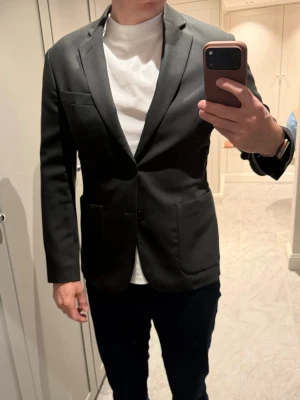Brun grön kavaj från J.Lindeberg - Säljer en stilren brun grön kavaj med slimmad passform och snygga detaljer som knappar på ärmsluten och två framfickor. Kavajen har en bröstficka och är tillverkad i ett material med struktur som ger ett elegant och intressant intryck. Perfekt för dig som vill ha en tidlös look. Modell: Hopper