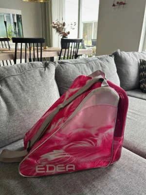 Rosa skridskoväska från Edea - Säljer en snygg rosa skridskoväska från Edea med glittriga detaljer och vita fjädermönster. Väskan har axelrem och bärhandtag, samt flera fack för smart förvaring. Perfekt för att bära skridskor och tillbehör till träningen.