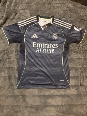 Real Madrid 25/26 Away Kit - Grå Real Madrid matchtröja från Adidas med vita detaljer och gula sömmar. Tröjan har klubbmärke och Adidas-logga på bröstet samt 'Emirates Fly Better' tryckt framtill. Slim fit-modell i lätt och ventilerande material, perfekt för fotbollsträning eller bara ett coolt utseende.