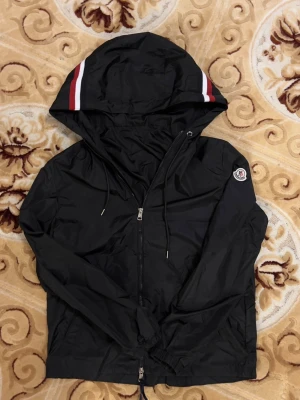 Svart Moncler vindjacka med huva - Svart vindjacka från Moncler med dragkedja, justerbar huva med röda och vita ränder samt logotyp på ärmen. Jackan har dragsko i midjan och är tillverkad i lätt, vattenavvisande material. Perfekt för en sportig och stilren look.