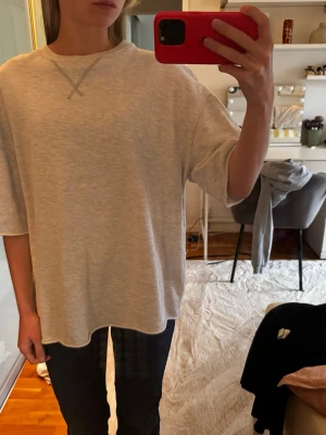 Oversized ljusgrå t-shirt - En oversized ljusgrå t-shirt med avslappnad passform och korta ärmar. T-shirten har en enkel design med en dekorativ söm vid halsringningen. Perfekt för en chill och stilren look.