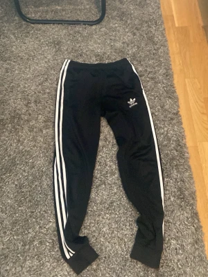 Svarta Adidas mjukisbyxor med vita ränder - Svarta mjukisbyxor från Adidas med klassiska vita ränder längs sidorna och logga på vänster lår. Byxorna har muddar vid bensluten och är i en smalare modell. Perfekta för en avslappnad och sportig stil.