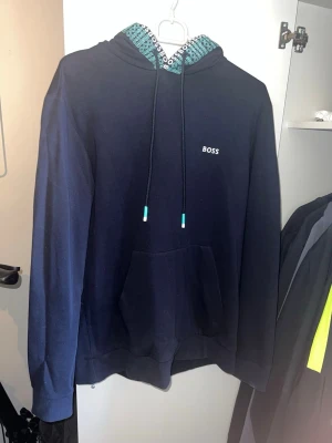 Hugo boss Hoodie  - Snygg mörkblå hoodie från BOSS med vit logga på bröstet och coolt mönstrat foder i huvan med BOSS-tryck i turkos och vitt. Klassisk känguruficka och dragsko med turkosa detaljer. Perfekt för en chill och stilren look.