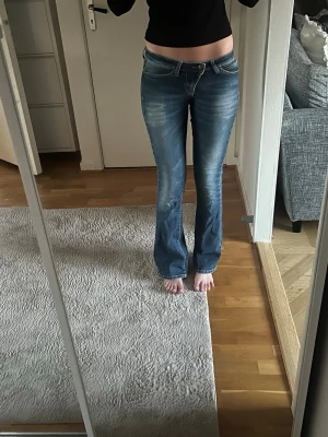 Blå bootcut jeans med slitningar - Snygga blå bootcut jeans med ljusa slitningar framtill och klassisk femficksdesign. Jeansen har låg midja och stängs med dragkedja och knapp. Perfekta för en avslappnad och trendig look.