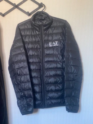 Svart dunjacka från EA7 Emporio Armani - Snygg svart dunjacka från EA7 Emporio Armani med diskret glansig finish och logga på bröstet. Jackan har ståkrage, dragkedja framtill och quiltad design. Perfekt för kyliga dagar och enkel att matcha med olika outfits.