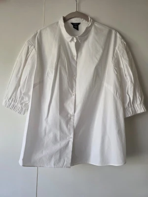 Vit kortärmad blus - Stilren vit blus från Lindex i bomull med trekvartsärmar med resår vid ärmslut. Klassisk krage och knäppning framtill. Perfekt för en clean och modern look.