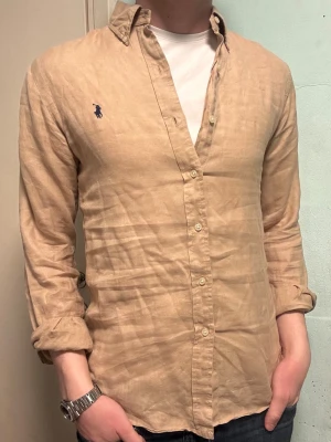 Beige Ralph Lauren linneskjorta – M Slim Fit - Säljer denna stilrena och snygga beiga linne Ralph Lauren skjorta. Skjortan är i mycket bra skick.  Perfekt nu inför sommaren.  Storlek S/P slim fit, men sitter  som en vanlig M slim fit.  Har du några frågor? Tveka inte att höra av dig. Allt gott från Deluxe garderoben ⭐️