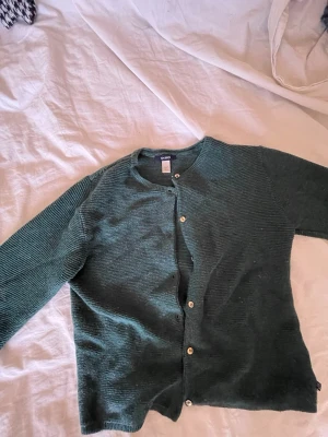 Grön ribbstickad kofta från Gina Tricot - Mysig grön kofta från Gina Tricot i ribbstickat material. Koftan har rund halsringning, långa ärmar och knäpps med ljusa knappar framtill. Perfekt att slänga på sig när det är lite kyligare ute.