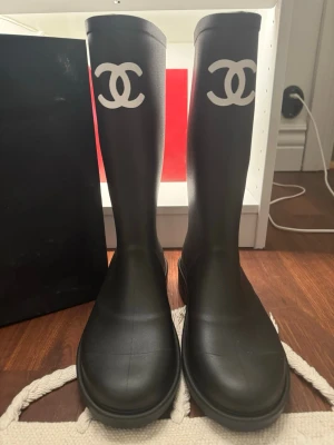 Svarta gummistövlar från Chanel - Snygga svarta gummistövlar från Chanel med det klassiska vita CC-logotypen framtill på skaftet. Stövlarna är höga och har en stilren, minimalistisk design i matt gummi. Perfekta för regniga dagar när du vill hålla stilen. Storlek 40. Mycket bra skick. Har påse till. 