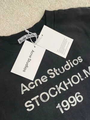 Svart långärmad tröja Acne Studios - Svart långärmad t-shirt från Acne Studios med vit text 'Acne Studios STOCKHOLM 1996' på bröstet. Tröjan har rund halsringning och är tillverkad i mjuk bomull. Perfekt för en stilren och trendig look.