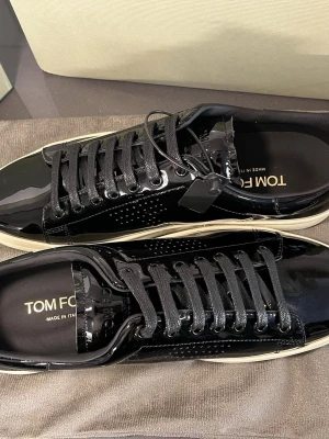  Tom Ford skor - Snygga svarta sneakers från Tom Ford i glansigt lackmaterial med svarta skosnören och vit sula. Klassisk låg modell med diskreta perforeringar på sidan och stilren design. Perfekta för dig som vill sticka ut med exklusiva detaljer.